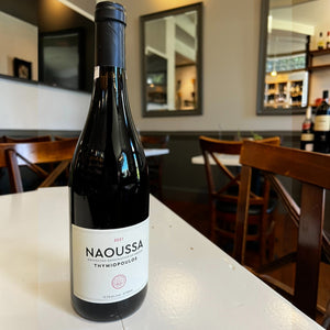 A bottle of THYMIOPOULOS ‘NAOUSSA’ XINOMAVRO 2021 red wine with a white label reading “2021 NAOUSSA PROTECTED DESIGNATION OF ORIGIN THYMIOPOULOS” and indicating 13.5% alcohol by volume.