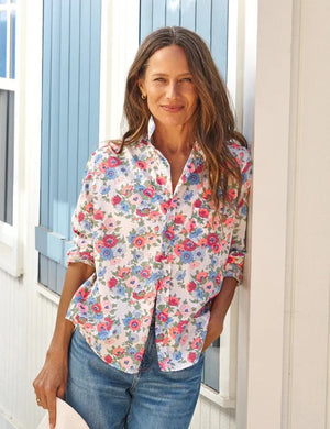 A woman with long brown hair stands smiling in front of a white wall with blue shutters, wearing the FRANK & EILEEN EILEEN Relaxed Button Up in Liberty Fabric—pink and red with blue flowers—paired with blue jeans.