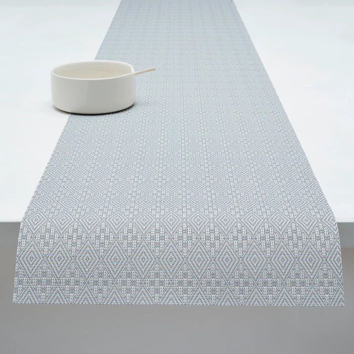 A white ceramic bowl with a spoon rests on the CHILEWICH - BOXWOOD TABLE MAT by CHILEWICH, showcasing a subtle geometric diamond pattern in light gray, set atop a white tabletop.