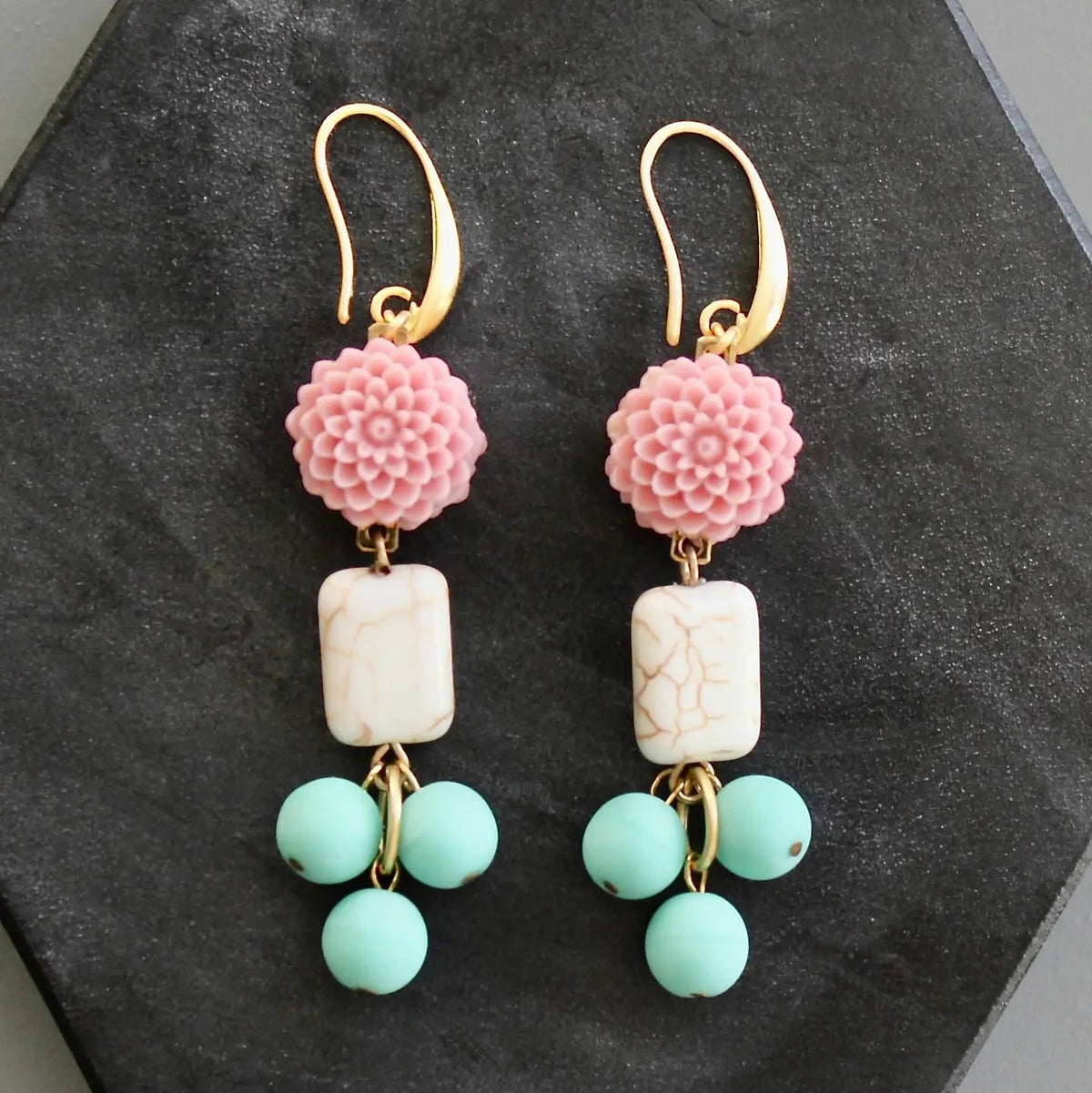 DAVID AUBREY - PINK AND TURQUOISE FLOWER EARRINGS by DAVID AUBREY feature gold hook fastenings, pink flower tops, rectangular white magnesite beads with a marble-like finish, and three dangling round mint-green Czech glass beads. Displayed on a dark hexagonal surface, these earrings blend delicate detailing with timeless elegance for a truly unique accessory.
