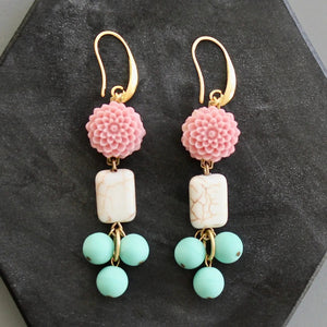 DAVID AUBREY - PINK AND TURQUOISE FLOWER EARRINGS by DAVID AUBREY feature gold hook fastenings, pink flower tops, rectangular white magnesite beads with a marble-like finish, and three dangling round mint-green Czech glass beads. Displayed on a dark hexagonal surface, these earrings blend delicate detailing with timeless elegance for a truly unique accessory.