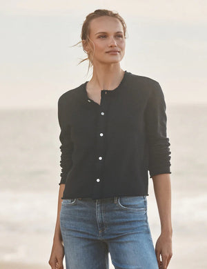 A woman with light brown hair stands outdoors by the beach, wearing FRANK & EILEEN’s JACKIE Sweatshirt Cardigan in British Royal Navy and blue jeans. She gazes into the distance with a relaxed expression; the background is softly blurred.
