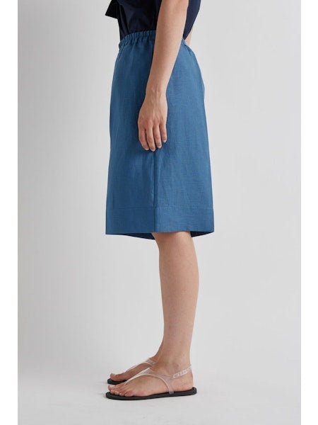 A woman stands against a plain background wearing the MEIMEIJ Pull On Linen Short in Cerulean, styled with a sleeveless navy wrap top, flat sandals, and her hair pulled back. Her expression is neutral.