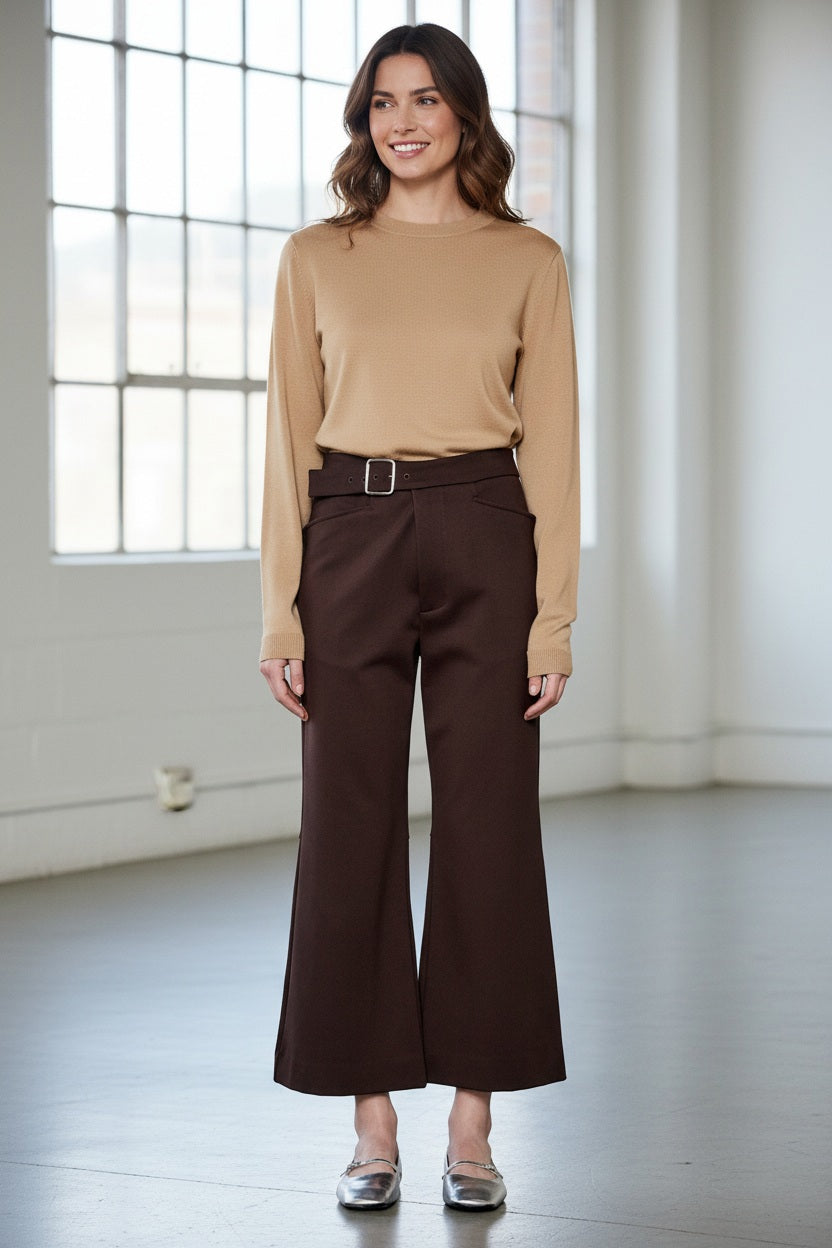 A person models the MEIMEIJ Crop Ponte Pant with Belt, shown in brown with pockets, paired with a beige top and silver flats. Only the lower half is visible against a plain white wall and concrete floor.