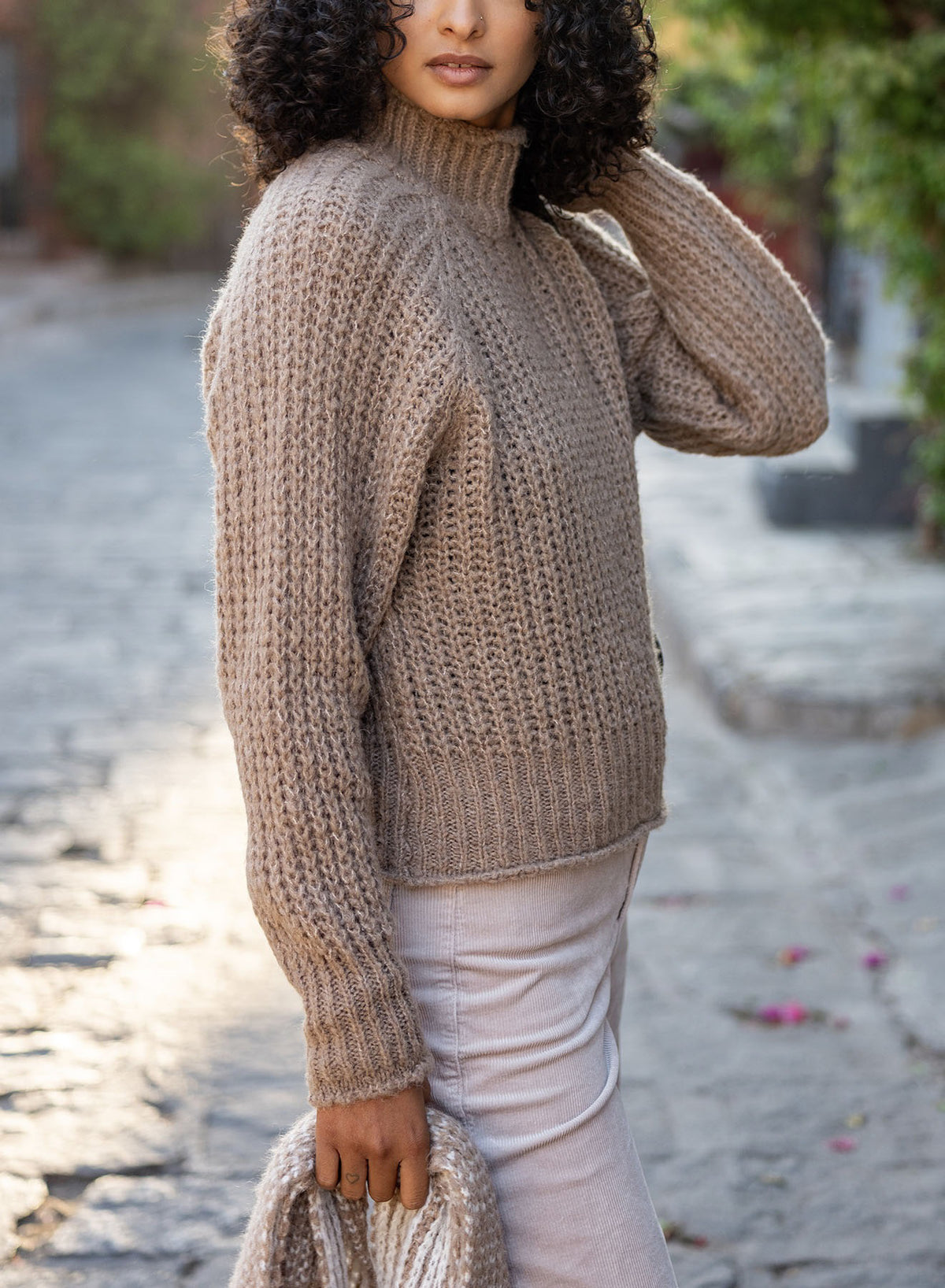 Wearing the MERSEA Beachside Softy Sweater in Beachstone, a person with curly hair stands on a cobblestone street holding a matching knit, the softly blurred background enhancing the cozy vibe.