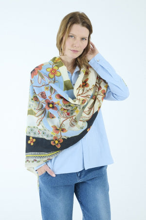 The STORIATIPIC ARIANE wool scarf with embroidery is adorned with a floral and vine pattern in red, green, blue, cream, and black tones. It features light blue edges and a soft, slightly fringed border. The embroidered scarf is loosely knotted against a white background.