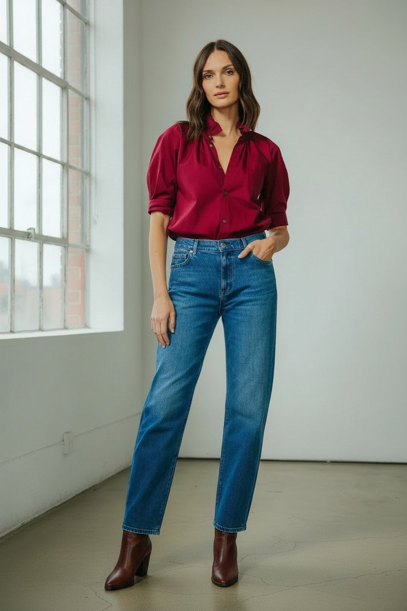 A woman stands indoors in front of a window, wearing the KMJ - SOUTHERN LOVER SHIRT IN BORDEAUX paired with blue jeans and brown boots, with one hand in her pocket.