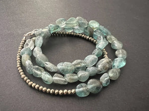 The ANN LIGHTFOOT - APATITE NUGGET TRIPLE WRAP BRACELET by ANN LIGHTFOOT features irregular light blue-green apatite stones, metallic round beads, and a sterling silver clasp, coiled in a circle on a dark surface.