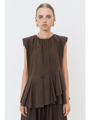 The individual is elegantly clad in the MEIMEIJ PLEATED ASYMETRICAL TOP, a sleeveless brown design with a unique peplum hem, paired with a matching long skirt. Gold hoop earrings and red flat shoes add flair against the plain white backdrop.