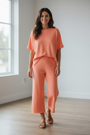 A woman stands outdoors on a stone patio, wearing FRANK & EILEEN'S Catherine Favorite Sweatpant in Wild Strawberry with a pink top and sandals. Steps, greenery, and a light-colored house are visible behind her.
