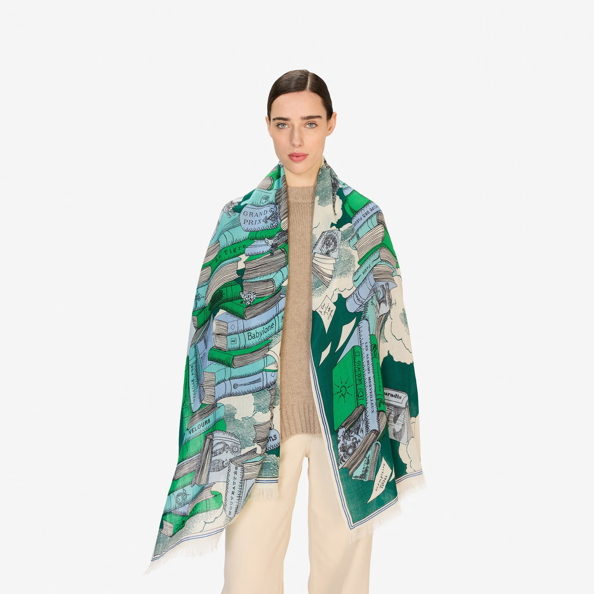 A woman with straight brown hair, in a beige sweater and cream pants, wears the INOUI EDITIONS Stack of Books Wool Scarf in Emerald, featuring green, blue, and white illustrated books and landscape designs.