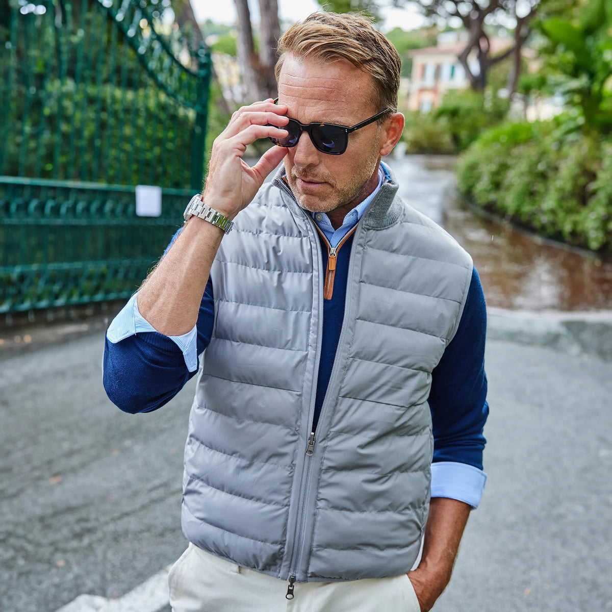 A smiling man in a light blue shirt and the COLLARS AND CO - REGENT REVERSIBLE QUILTED WOOL VEST holds a blue umbrella on a rainy street—showcasing COLLARS AND CO's ideal insulated layering for wet days.