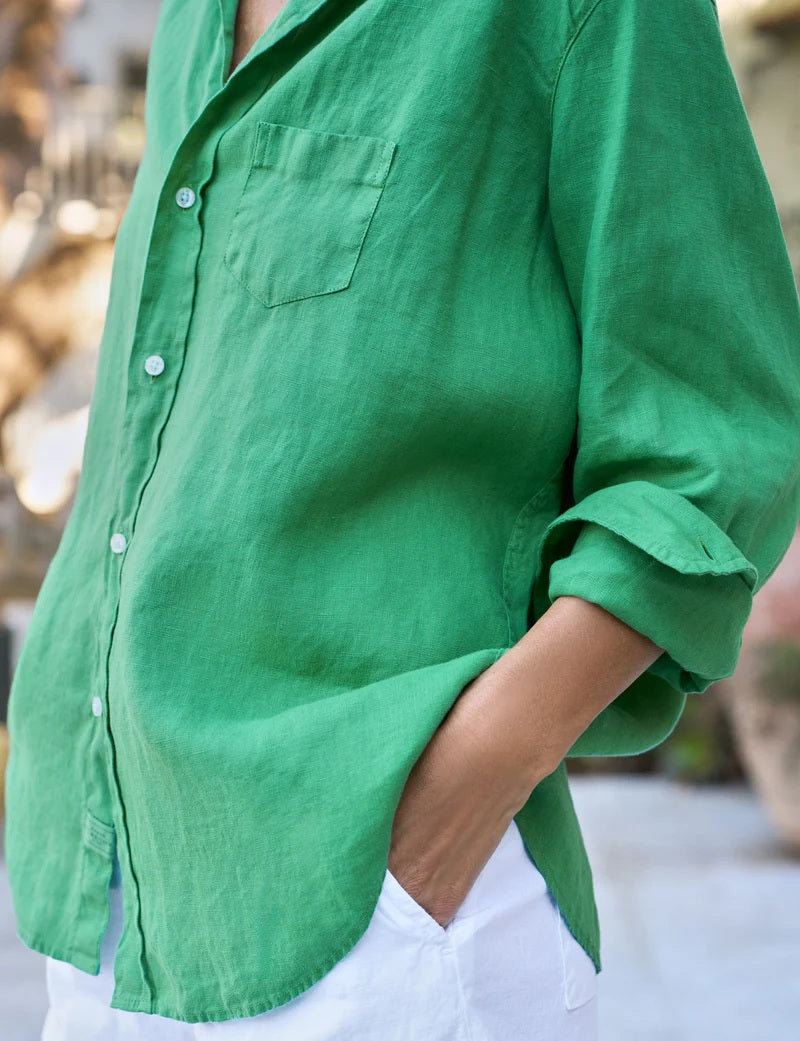 A woman with long brown hair smiles outdoors, wearing the FRANK &amp; EILEEN Eileen Relaxed Button Up in washed linen Kelly Green and white shorts. She stands with one hand in her pocket amid greenery and large potted plants.