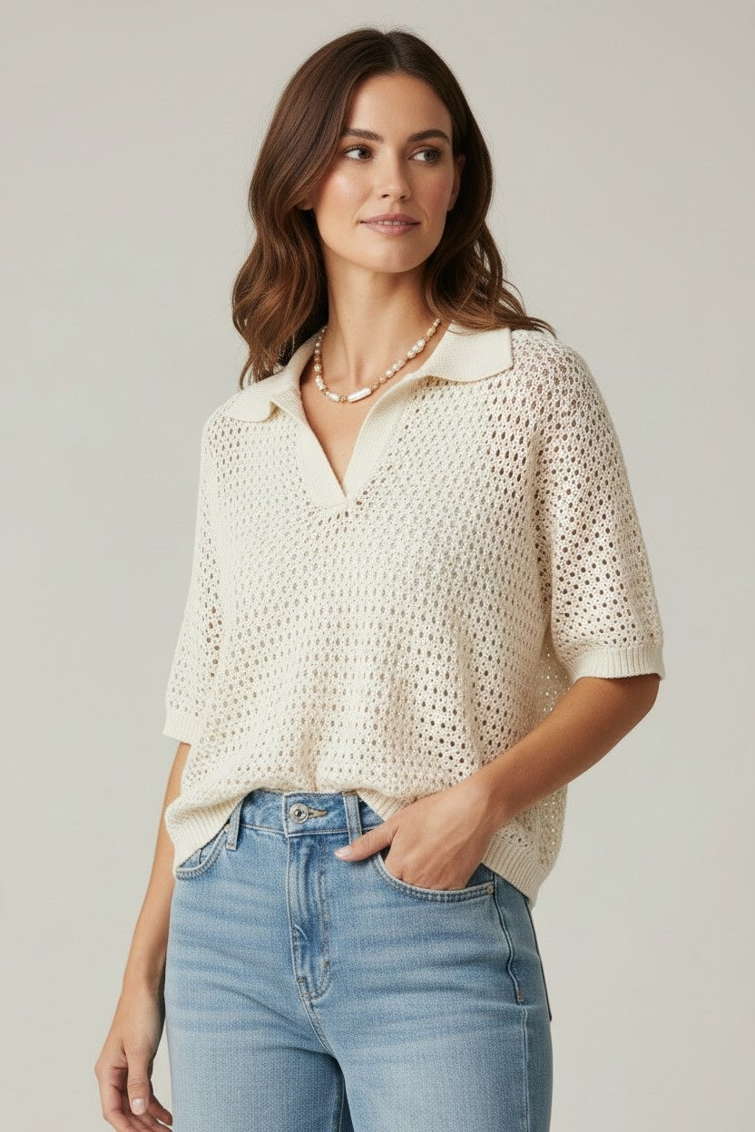 A woman stands against a plain background, wearing the BASE MILANO Short Sleeve Splitneck Cotton Polo in Ivory with light blue high-waisted wide-leg jeans, beige shoes, and a necklace. She has brown wavy hair and poses with one hand in her pocket for a chic BASE MILANO-inspired look.