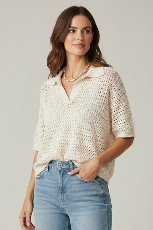 A woman stands against a plain background, wearing the BASE MILANO Short Sleeve Splitneck Cotton Polo in Ivory with light blue high-waisted wide-leg jeans, beige shoes, and a necklace. She has brown wavy hair and poses with one hand in her pocket for a chic BASE MILANO-inspired look.