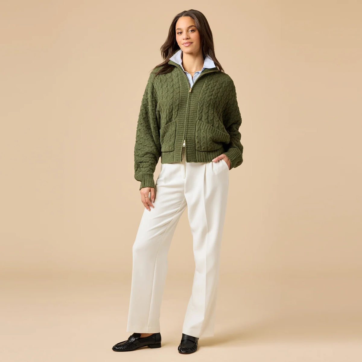 A woman wearing a cream-colored, plush SOFTIES - CABLE KNIT MARSHMALLOW FUNNEL NECK DOUBLE ZIP JACKET with a funnel neck and double zipper, paired with blue jeans.