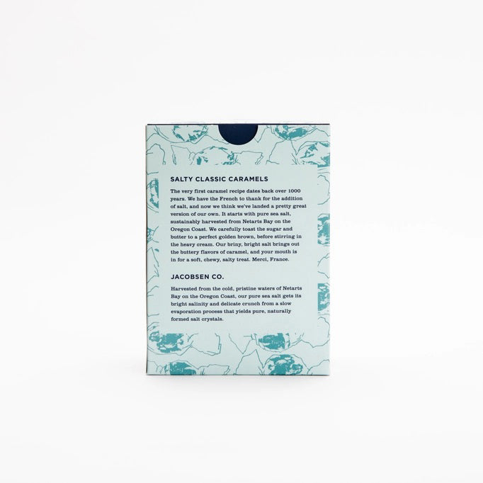 A light blue box of JACOBSEN SALT CO. JACOBSEN SALT - SALTY CARAMELS BOX, featuring a patterned design and product label on the front, rests on a light surface.