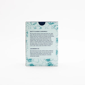 A light blue box of JACOBSEN SALT CO. JACOBSEN SALT - SALTY CARAMELS BOX, featuring a patterned design and product label on the front, rests on a light surface.