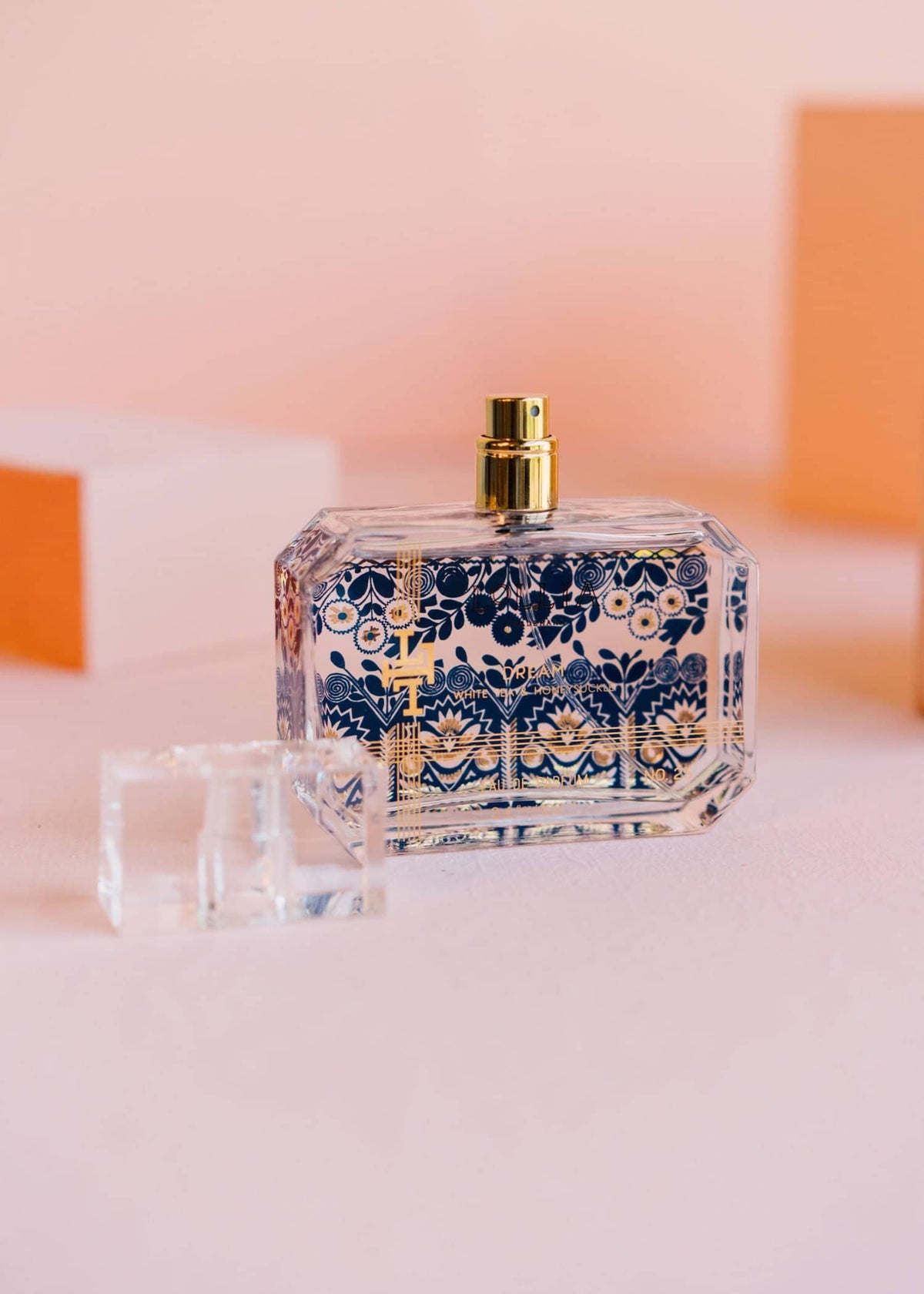 A rectangular perfume bottle with intricate blue patterns and gold accents lays on a pink surface, leaning against a light pink block. The elegant bottle, labeled DREAM EAU DE PARFUM by MARGOT ELENA, features a clear cap that allows visibility of the liquid inside.