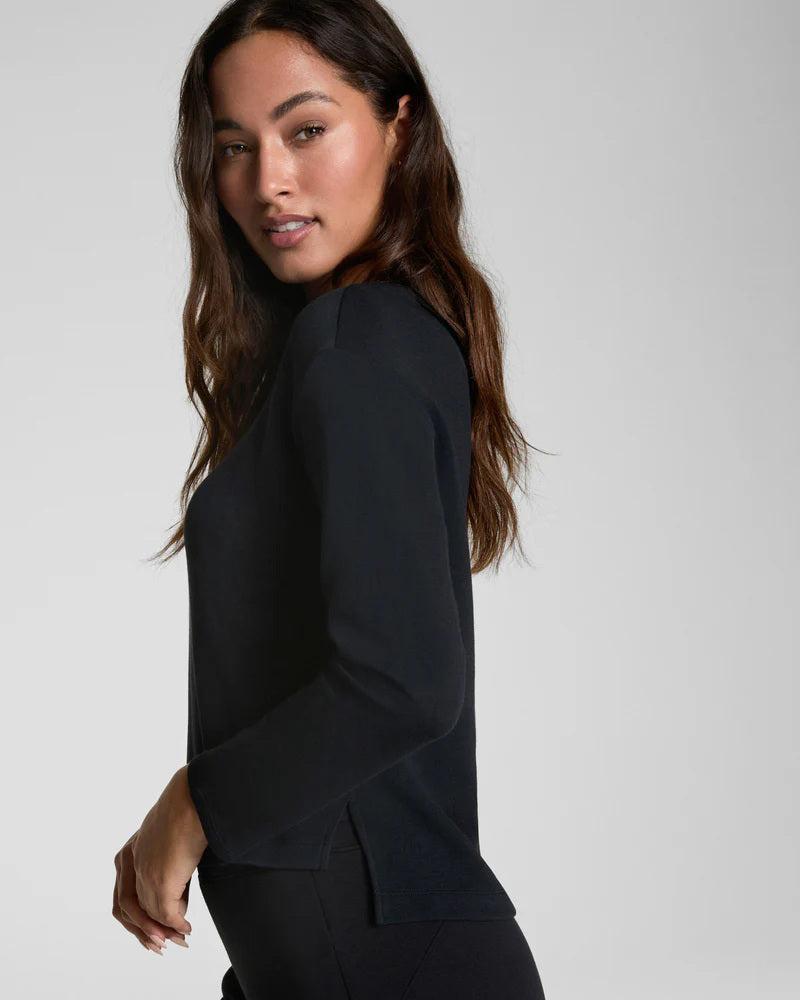 A woman with long dark hair wears the SPANX AirEssentials Relaxed Long Sleeve Top and matching black pants, standing against a plain light gray background and looking forward with a neutral expression.