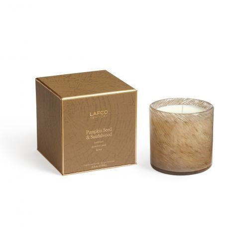 A candle in a transparent glass container, featuring a rich brown hue, is placed beside a coordinating brown box adorned with gold details. The set exudes a gourmand aroma and is elegantly labeled as &quot;LAFCO - Pumpkin Seed &amp; Sandalwood Candle 6.5oz.&quot; The candle&#39;s texture appears soft like fur.