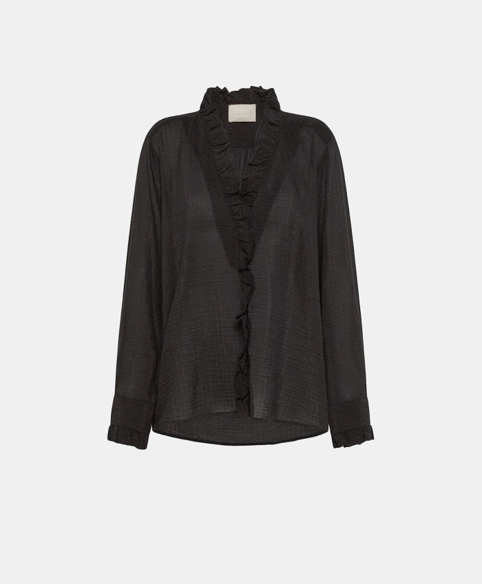 A person with shoulder-length blonde hair is wearing the MOMONI - Marc Blouse in textured cotton silk, which features ruffles along the collar and cuffs. They are also wearing black wide-leg pants and holding a teal-colored clutch. The background includes a light-colored door and wall.