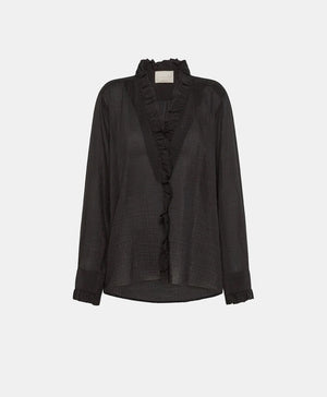 A person with shoulder-length blonde hair is wearing the MOMONI - Marc Blouse in textured cotton silk, which features ruffles along the collar and cuffs. They are also wearing black wide-leg pants and holding a teal-colored clutch. The background includes a light-colored door and wall.