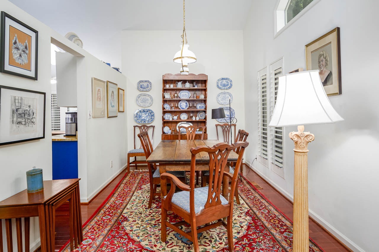 A traditional dining room with a wooden table and six chairs, a red patterned rug, a tall lamp, framed artwork on the walls, and a wooden hutch displaying blue and white plates at the far end.