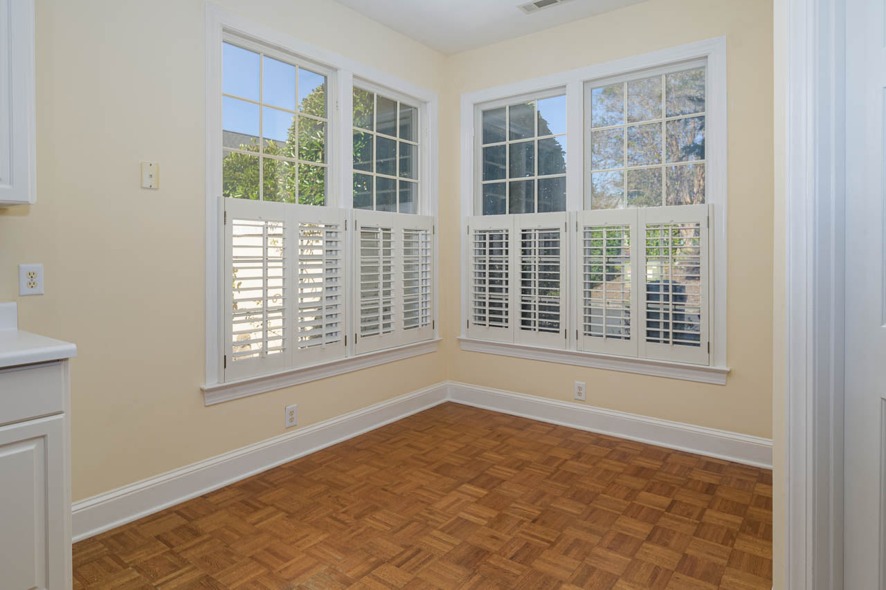 Sunlit corner of a room with tan walls, wood parquet flooring, and large windows with white shutters; trees and outdoor scenery visible through the windows.
