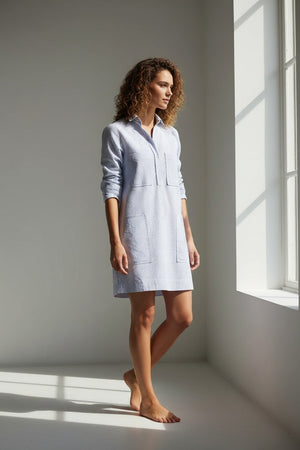 A woman wearing the FRANK & EILEEN Southern Ireland Utility Dress in Italian Performance Linen Navy and White Stripe, designed with six pockets, strolls barefoot along a white pathway by the ocean near a lighthouse, surrounded by flowers, palm trees, and a bird on a sunny day.