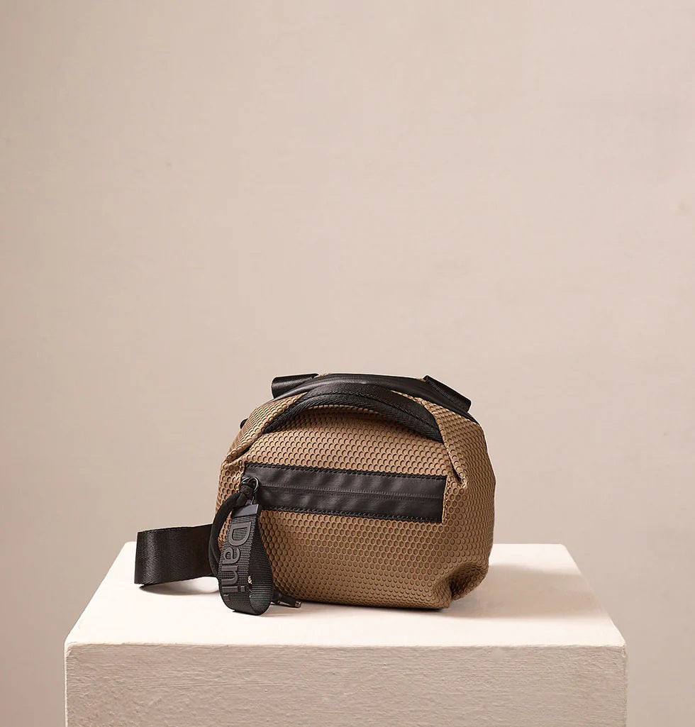 The DANIELLA LEHAVI BONNIE MINI TRAVEL CROSSBODY, a small brown vegan bag with black accents and strap, is displayed on a white pedestal against a plain beige background.
