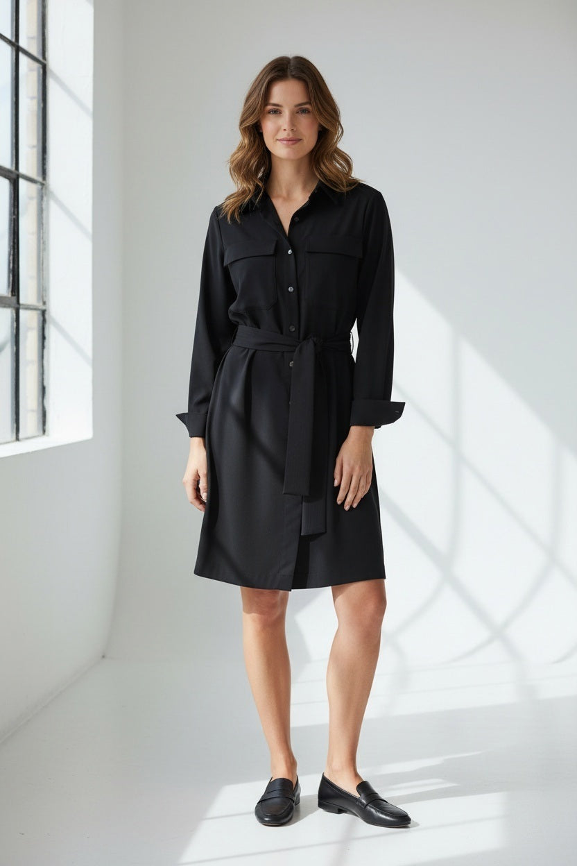 A woman stands facing forward, wearing a long-sleeved, button-up black shirt dress with a tied belt at the waist. She has long brown hair and neutral expression, set against a plain light background.