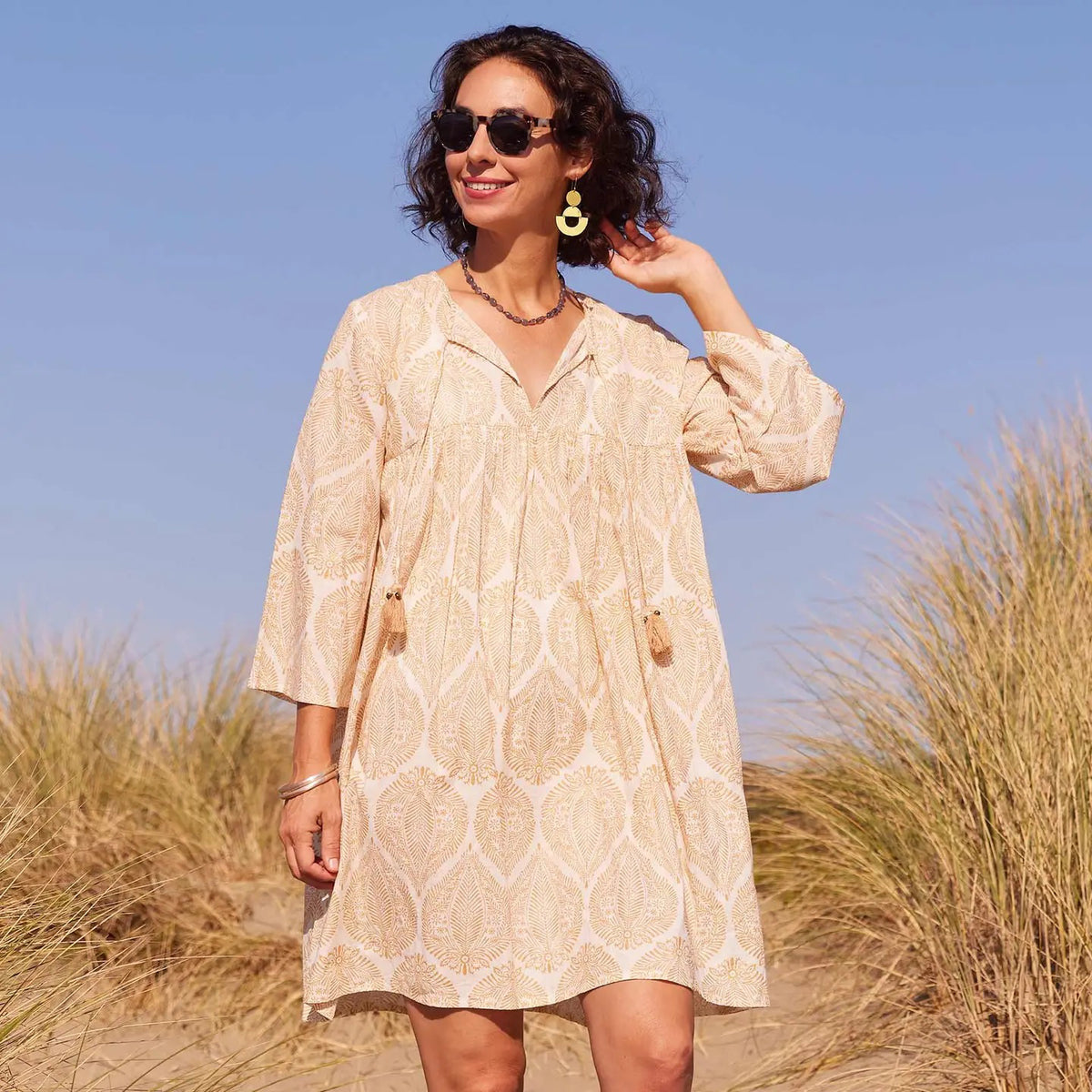 A woman smiles and looks down while wearing the ROCKFLOWERPAPER - ZAHARRA DRESS, a lightweight cream long-sleeve coverup with a subtle pattern and tassels, standing against a beige background.