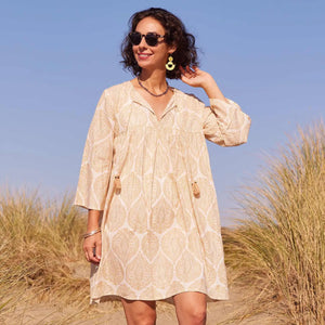 A woman smiles and looks down while wearing the ROCKFLOWERPAPER - ZAHARRA DRESS, a lightweight cream long-sleeve coverup with a subtle pattern and tassels, standing against a beige background.