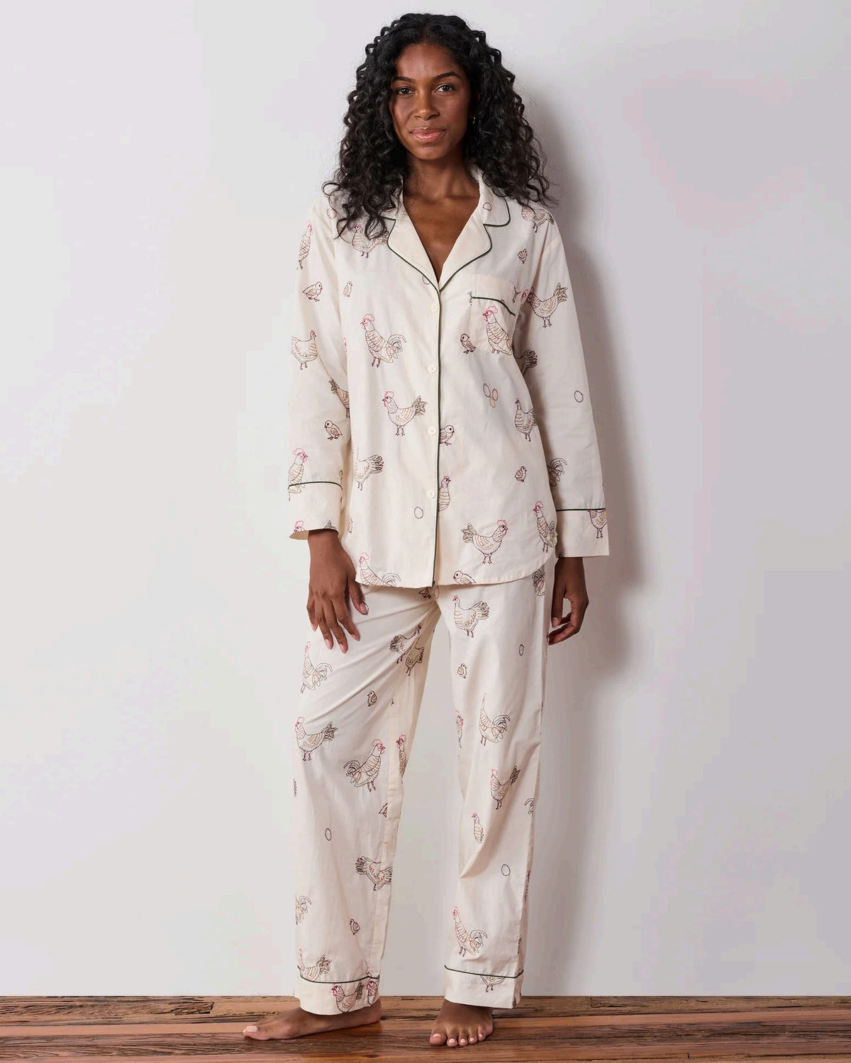 A woman dressed in the PRINTFRESH Embroidered Hens Long Pajama Set in Buff smiles as she stands by a wooden fence, holding a basket and watching chickens peck at the ground in a farmyard setting, with trees and a path visible behind her.
