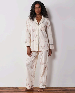 A woman dressed in the PRINTFRESH Embroidered Hens Long Pajama Set in Buff smiles as she stands by a wooden fence, holding a basket and watching chickens peck at the ground in a farmyard setting, with trees and a path visible behind her.