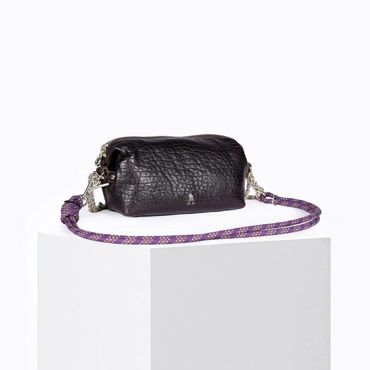 The CRAIE STUDIO - NUAGE BUBBLE POURPRE MINI BAG, a compact and textured black lambskin accessory featuring silver hardware and an eye-catching purple patterned strap, is showcased on a white pedestal against a plain background.