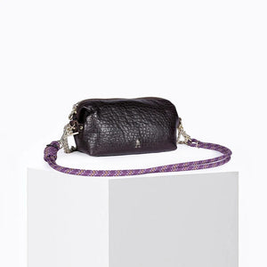 The CRAIE STUDIO - NUAGE BUBBLE POURPRE MINI BAG, a compact and textured black lambskin accessory featuring silver hardware and an eye-catching purple patterned strap, is showcased on a white pedestal against a plain background.