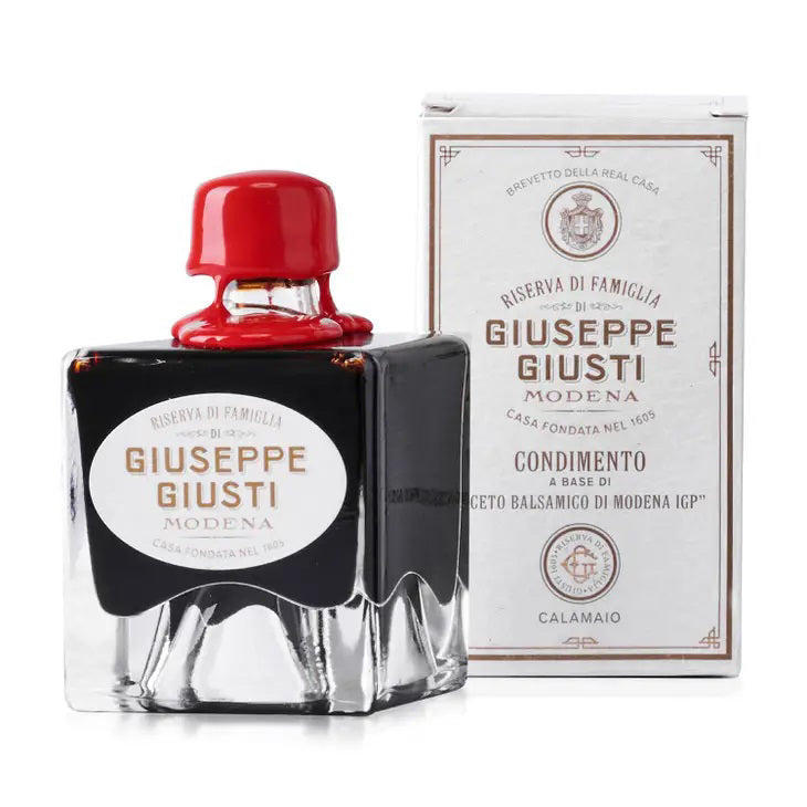 A square glass bottle with a red wax seal cap containing Mercato di Bellina's Giuseppe Giusti - Family Reserve Balsamic Vinegar stands next to a white box with product information. The bottle features a black label with gold detailing, and the box displays similar branding and text.