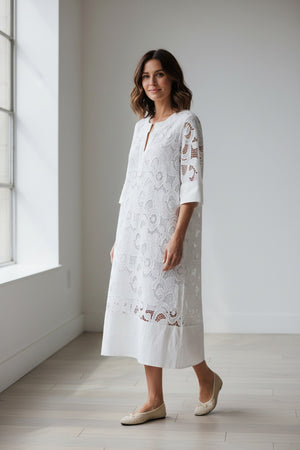 A woman wears the MILVA MI - EMBROIDERED COTTON DRESS IN WHITE by MILVA, styled with bold black and white earrings and black sunglasses. Her straight blonde hair and hand on her hip complete a chic summer resort look.