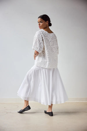A woman stands in profile wearing the HOPE FOR FLOWERS EYELET TEE, a white, short-sleeved textured lace top made from GOTS certified organic cotton, paired with a flowing white skirt. Her hair is pulled back with a plain light background.