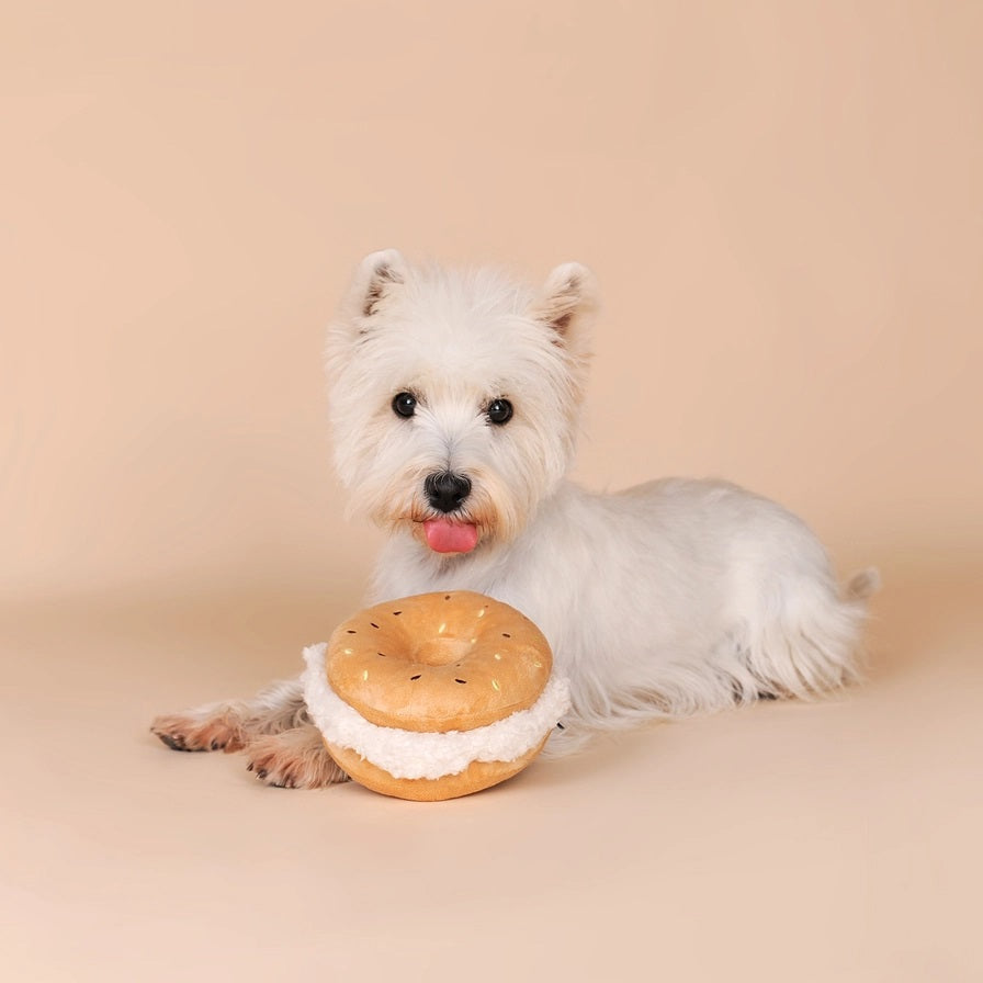 A small, fluffy white dog sniffs the CHEWFFON BAGEL PLUSH SQUEAKY DOG TOY, a large, round, beige plush toy resembling a sandwich, set against a plain light beige background.
