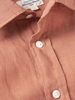 A person wears the HARTFORD - PAUL LINEN SHIRT IN FADED COPPER by HARTFORD, paired with light pants, standing with one hand in their pocket against a plain background.
