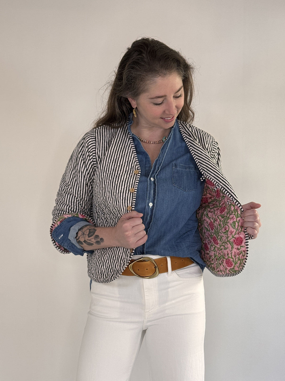 A woman with visible forearm tattoos smiles slightly against a plain background, wearing the FARYLINA ZOIE Reversible Quilted Jacket over a denim shirt, white pants, and a tan belt.