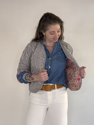 A woman with visible forearm tattoos smiles slightly against a plain background, wearing the FARYLINA ZOIE Reversible Quilted Jacket over a denim shirt, white pants, and a tan belt.