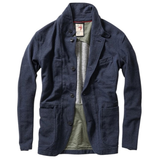 A person wearing the RELWEN - PIQUE TRAP BLAZER IN NAVY HEATHER over a checked shirt and green cargo pants stands with hands in pockets against a plain background, showcasing the blazer’s relaxed fit.