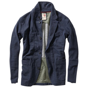 A person wearing the RELWEN - PIQUE TRAP BLAZER IN NAVY HEATHER over a checked shirt and green cargo pants stands with hands in pockets against a plain background, showcasing the blazer’s relaxed fit.