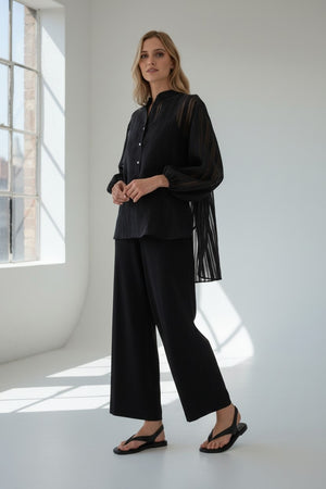 A woman stands in a bright room, looking at the camera with a neutral expression. She is wearing the VICARIO CINQUE SEAN SHEER STRIPE SHIRT, a black sheer long-sleeve blouse crafted from lightweight fabric with a band collar by VICARIO CINQUE, paired with black wide-leg pants and black sandals.