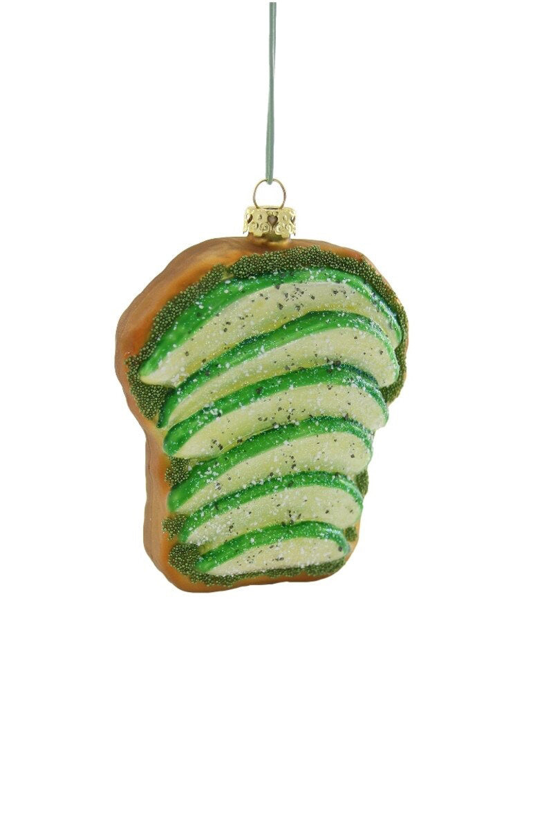 The AVOCADO TOAST ORNAMENT by CODY FOSTER is a decorative glass piece shaped like toast topped with avocado slices, designed with glitter and suspended from a string. It features a glossy finish that artfully combines green and brown hues, while its transparency enhances its elegant appeal.