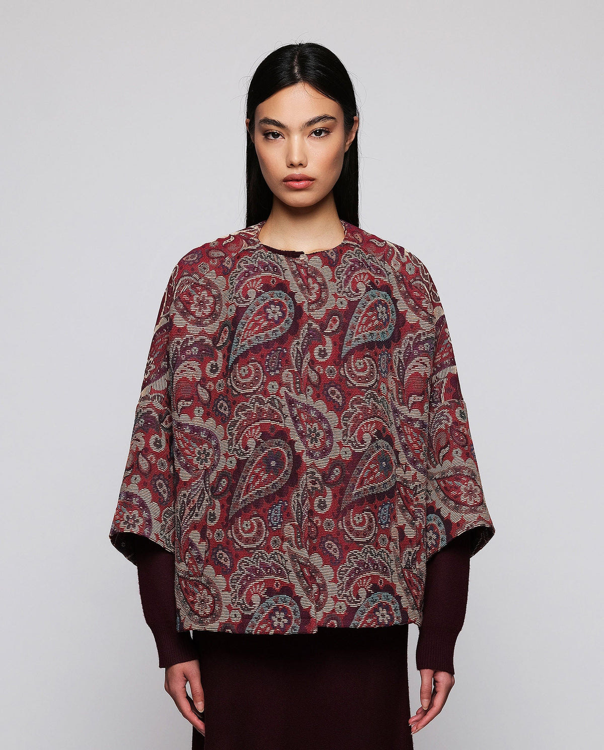 A woman stands against a plain background wearing the MIRTO 1956 Burgundy Paisley Jacquard Jacket—an oversized, cotton-blend piece in rich red and purple tones from MIRTO 1956—styled over a long burgundy dress and black shoes. The relaxed fit of the jacket complements her straight dark hair and neutral expression.